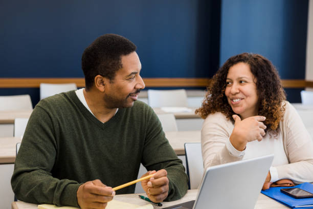 The couple smiles and talks together before their evening class.  He has his laptop open and ready and she has her smart phone on the desk in front of her.